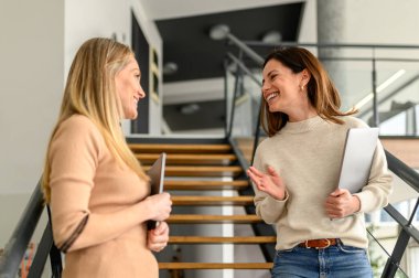 Ofisteki merdivenlerde dururken dizüstü bilgisayarlarıyla gülümseyen ve tartışan kadın profesyonellerin düşük açılı görüntüsü.