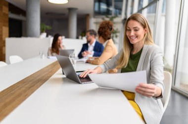 Kadın avukat, arka planda meslektaşlarıyla konferans odasında çalışırken belgeleri analiz ediyor ve laptop kullanıyor.