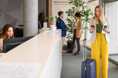 Bavulu ve pasaportu olan kendine güvenen bir iş kadını otel resepsiyonunda beklerken akıllı telefon kullanıyor.