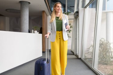 Bavulu ve pasaportu olan, pencereden bakan ve modern bir otelde yürüyen kendine güvenen bir iş kadını portresi.