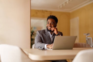 Düşünceli genç işadamı, modern ofisteki masada bilgisayarla çalışırken başka tarafa bakıyor.
