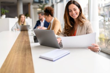 Modern ofiste arka planda iş arkadaşlarıyla bilgisayarla çalışırken hukuki belgeleri analiz ederken gülümseyen bir profesyonel.