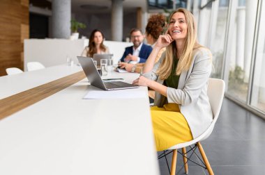 İş arkadaşları arka planda tartışırken, gülümseyen kadın yönetici bilgisayarda çalışıyor.