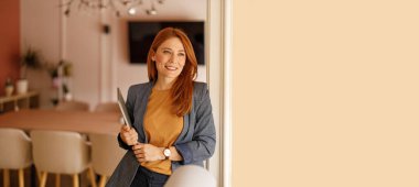 Dizüstü bilgisayarı olan mutlu bir kadın muhasebeci ofiste duvarın yanında dururken başka yöne bakıyor ve stratejiler düşünüyor.