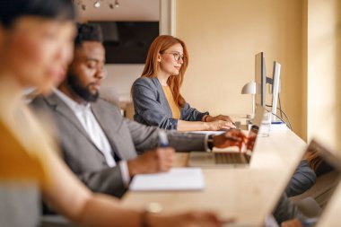 Gözlüklü odaklanmış iş kadını, modern ofiste çalışma masasında meslektaşlarıyla otururken bilgisayar başında çalışıyor.