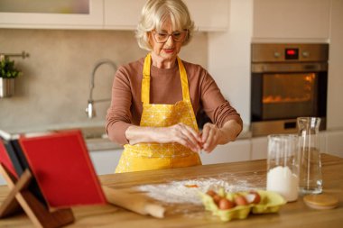 Önlüklü, kendine güveni tam yaşlı bir kadın mutfak adasında un kırarken çevrimiçi blog için şekerleme yapıyor.