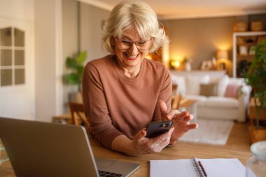 Masasında dizüstü bilgisayarı olan yaşlı bir kadın evdeki sanal sağlık kontrolü için doktorla iletişime geçmek için cep telefonu kullanıyor.