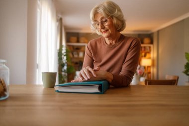 Sarı saçlı ve elleri kapalı aile albümü olan düşünceli son sınıf öğrencisi kadın evdeki ahşap yemek masasında oturuyor.