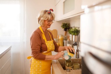 Önlüklü ve gözlüklü yaşlı kadın mutfak lavabosunda günlük ev işleri yapıyor.