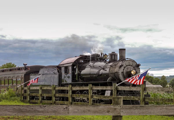 曇った日にはアメリカ国旗が風になびくフェンスの上に 蒸気機関車が近づいてくる — ストック写真