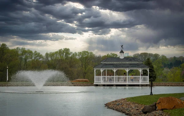 stock image View of a Fountain and Large Gazebo on a Pond With Clouds and The Sun Breaking Thru on a Spring Day