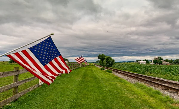 stock image On a Cloudy Day American Flags are Waving in the Wind, on a Fence near a Single Railroad Track