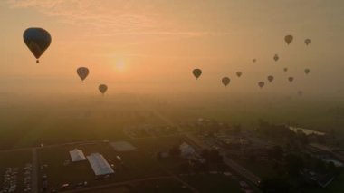 An Aerial View of Multiple Hot Air Balloons Floating into the Early Mist With a Red Sun During a Festival With Crowds Watching, on a Summer Day