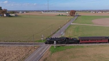 Ronks, Pennsylvania, November 4, 2022 - Aerial Parallel View of a Steam Passenger Train on a Single Track Thru Farmlands Approaching a Crossing on a Sunny Day