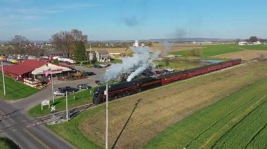 Ronks, Pennsylvania, November 9, 2022 - Drone Ahead View of a Steam Passenger Train Blowing Lots of Smoke and Steam and Crossing a Rail Road Crossing on a Sunny Fall Day