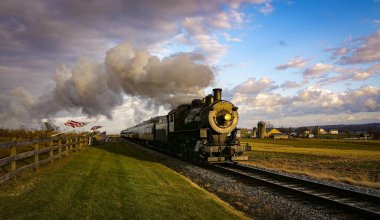 A View of a Classic Steam Passenger Train Approaching, With American Flags Attached to a Fence on a Sunny Autumn Day