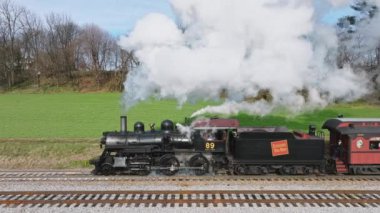 Ronks, Pennsylvania, December 4, 2022 - An Aerial Parallel View of a Antique Steam Passenger Train, Starting to Slowly Pull out of a Station, Puffing Lots of Smoke, on a Sunny Winter's Day