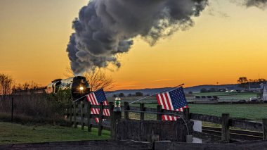A View of an Antique Stream Passenger Train, Traveling at Sunrise, While passing a Fence with American Flags on it, on an Autumn Day