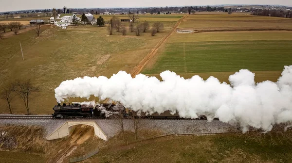 stock image A Side Aerial View of a Steam Passenger Train Approaching, Traveling Thru Open Farmlands, Blowing Lots of White Smoke, on a Winter Day