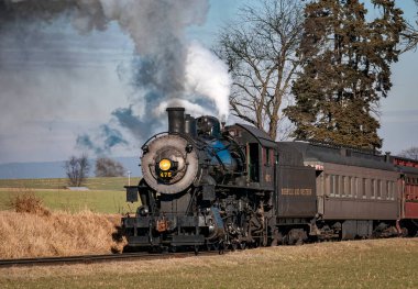 Ronks, Pennsylvania, December 30, 2022 - A View of a Classic Steam Passenger Train Approaching, Traveling Thru the Countryside, Blowing Smoke and Steam on a Winter Day