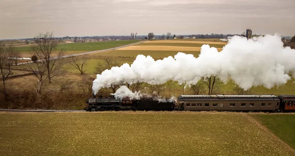 stock image A Side Aerial View of a Steam Passenger Train Approaching, Traveling Thru Open Farmlands, Blowing Lots of White Smoke, on a Winter Day