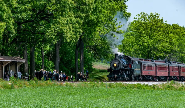 stock image Ronks, Pennsylvania, May 18, 2023 - A View of an Antique Passenger Steam Train Approaching a Small Station To Pick Up a Group of Passengers on a Sunny Day