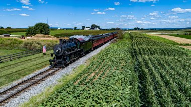 Güneşli bir yaz gününde Buharlı Yolcu Treni 'ne yaklaşan, Thru Tarım Alanlarını gezen ve Amerika Bayrağıyla Çitleri aşan İnsansız Hava Aracı.