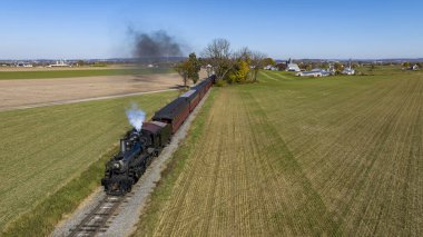 Buharlı Yolcu Treni 'nin Önünden ve Yukarıdan Bir İHA Görünümü Tarım Alanlarında Seyahat Ediyor ve Güneşli Sonbahar Günü Duman Esiyor