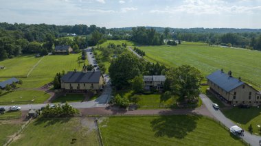 Yeşil tarlalarla çevrili, ağaçlarla döşenmiş, sakin bir kırsal alan manzarası ve konut ve çiftlik binaları koleksiyonuyla çevrili bir ana yol.