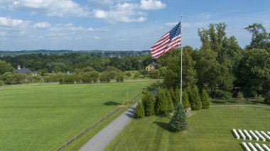 Uçsuz bucaksız, sarmal bir yol ve ahşap çitlerle çevrili geniş yeşil bir alan, önde gelen bir Amerikan bayrağı dalgalanan, dağılmış bulutların altındaki binalar ve ağaçlarla süslenmiş kırsal bir araziye bakan,.
