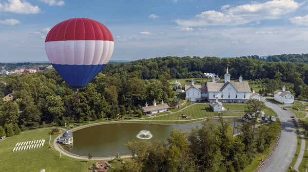 Vibrant Hot Air Balloon With Red, White, And Blue Stripes And Large Letters Floating In The Sky Above A Scenic Landscape Featuring Orthodox Church Buildings, A Reflective Pond, And Manicured Lawns.