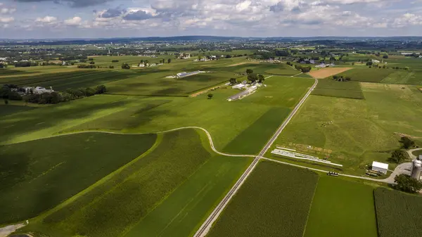 stock image Expansive Aerial View Of Rolling Green Agricultural Fields, Farm Buildings, And A Long Country Road Under A Cloudy Sky.