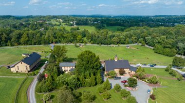 Ağaçların ve ağaçların oluşturduğu uzak ufuk ve bulutlu gökyüzünün altındaki tepelerin oluşturduğu geniş yeşil çitlerin ortasındaki binalara giden kıvrımlı bir yol üzerindeki hava manzarası..