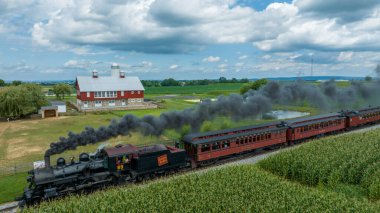 Ronks, Pennsylvania, 15 Ağustos 2023 - harmanlanmış bir hareketsizlik ve durgunluk içinde, bir buhar treni sakin bir çiftliğin yanında tıkırdıyor, kırmızı bir ahır ve yeşillik tarlaları ile birlikte..