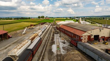 Strasburg, Pennsylvania, August 15, 2023 - An expansive view from above captures a rural rail yard with freight cars, surrounded by the lush, orderly farmlands stretching into the distance. clipart