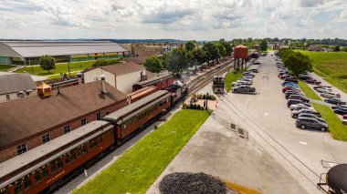 Strasburg, Pennsylvania, 15 Ağustos 2023 - Büyüleyici bir tren istasyonundan kalkmak üzere olan eski bir buhar treni, günümüzün modern araba ve yeşillik zeminine karşı eski bir dönemi anımsatıyor..