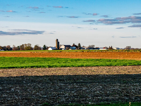 The cool dusk light washes over sprawling farmlands, highlighting the rich tapestry of tilled earth and burgeoning crops with a farmstead silhouette in the distance.
