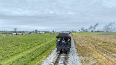 Ronks, Pennsylvania, ABD, 27 Nisan 2024 - Trenin arkasından çekilen sakin bir yolculuk, yolcuların bulutlu bir gökyüzü altında, çiftlik evleri ve yemyeşil tarlaları olan, kırsal alanda gezinmelerini gösteriyor..