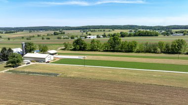 Kırsal bir çiftliğin huzurlu güzelliğini gösteren geniş bir hava görüntüsü, açık mavi gökyüzü altında sürülen tarlaların, yeşil ekinlerin ve çiftlik binalarının karışımını gösteriyor..