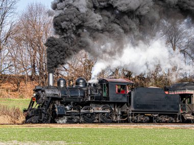 Bir buhar treni raylarda ilerliyor, arkasında bir duman izi bırakıyor. Tren siyah ve kırmızı bir kapısı var. Duman yoğun ve dalgalı, hareket ve güç hissi yaratıyor.