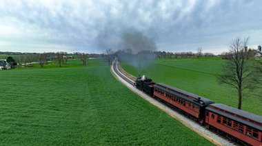 Etkileyici bir hava fotoğrafı. Yoğun bir duman bulutu yayan tarihi bir buhar treni. Çarpıcı bir bulutlu gökyüzü altında canlı yeşil alanlar arasında süzülüyor..