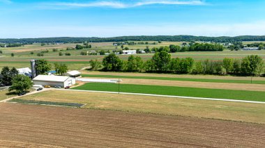 Kırsal bir çiftliğin huzurlu güzelliğini gösteren geniş bir hava görüntüsü, açık mavi gökyüzü altında sürülen tarlaların, yeşil ekinlerin ve çiftlik binalarının karışımını gösteriyor..