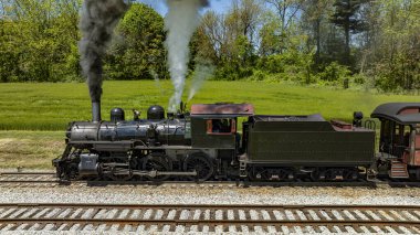 89 numaralı vintage buharlı lokomotifin detaylı bir görüntüsü. Parlak, güneşli bir günde yemyeşil bir alanda çalışırken yoğun duman ve buhar yayan motoru gösteriyor..