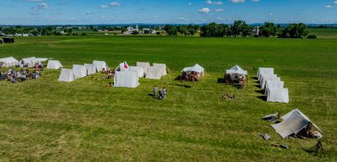 Tarlaları çadır olan ve yeşil bir alanda yeşil giysiler giyen, arka planda çiftlik binaları olan kırsal manzaralı, çadır çadırları ve katılımcılarla dolu tarihi bir yeniden canlandırma kampı manzarası..