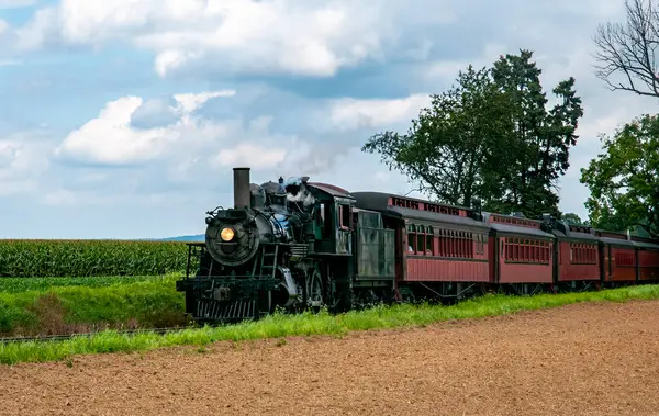 89 numaralı siyah buharlı lokomotif Strasburg isimli kırmızı yolcu arabalarını çekiyor yemyeşil ekinler ve ağaçlarla dolu kırsal bir alanda bulutlu mavi gökyüzü altında seyahat ediyor..