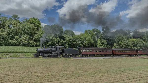 stock image A Black Steam Locomotive Pulling Red Passenger Cars Passes Through A Verdant Countryside With Lush Green Trees In The Background, Billowing Smoke Against A Partly Cloudy Sky.