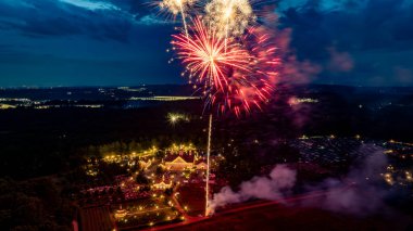 Geceleyin, ağaçlar ve tarlalarla çevrili, parlak gökyüzü ve uzak şehir ışıkları ile aydınlık bir arazide patlayan renkli havai fişekler bir kutlama atmosferini yakalıyor..
