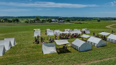 Beyaz tuval çadırlı tarihi bir yeniden canlandırma kampı manzarası, periyot giysili insanlar, ve bayraklar, yeşil bir alana kurulmuş çiftlik binaları ve açık güneşli bir alanda yuvarlanan tepeler.