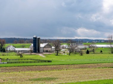 Ronks, Pensilvanya, ABD, 4 Nisan 2024 - Yeşil Alanlar ve Yuvarlanan Tepeler 'de, Bulutlu Gökyüzü Altında Çevrelenmiş Beyaz Binaları, Ahırları ve İki Uzun Silosu olan Huzurlu Bir Çiftlik