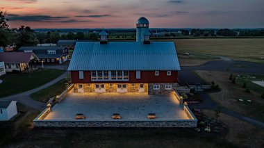Ronks, Pennsylvania, ABD, 19 Temmuz 2024 - Alacakaranlıkta Sakin Çiftlik Sahnesi Beyaz Silolu Büyük Kırmızı Ahır, Aydınlanmış Taş Avlu, Yakın Bir Tuğla Evi ve Genişletilmiş Yeşil Tarlalar